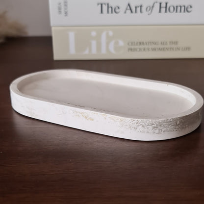 Side view of a natural white oval trinket tray with light gold metal streaks on wood surface. In the background 'The Art of Home' and 'Life' books.