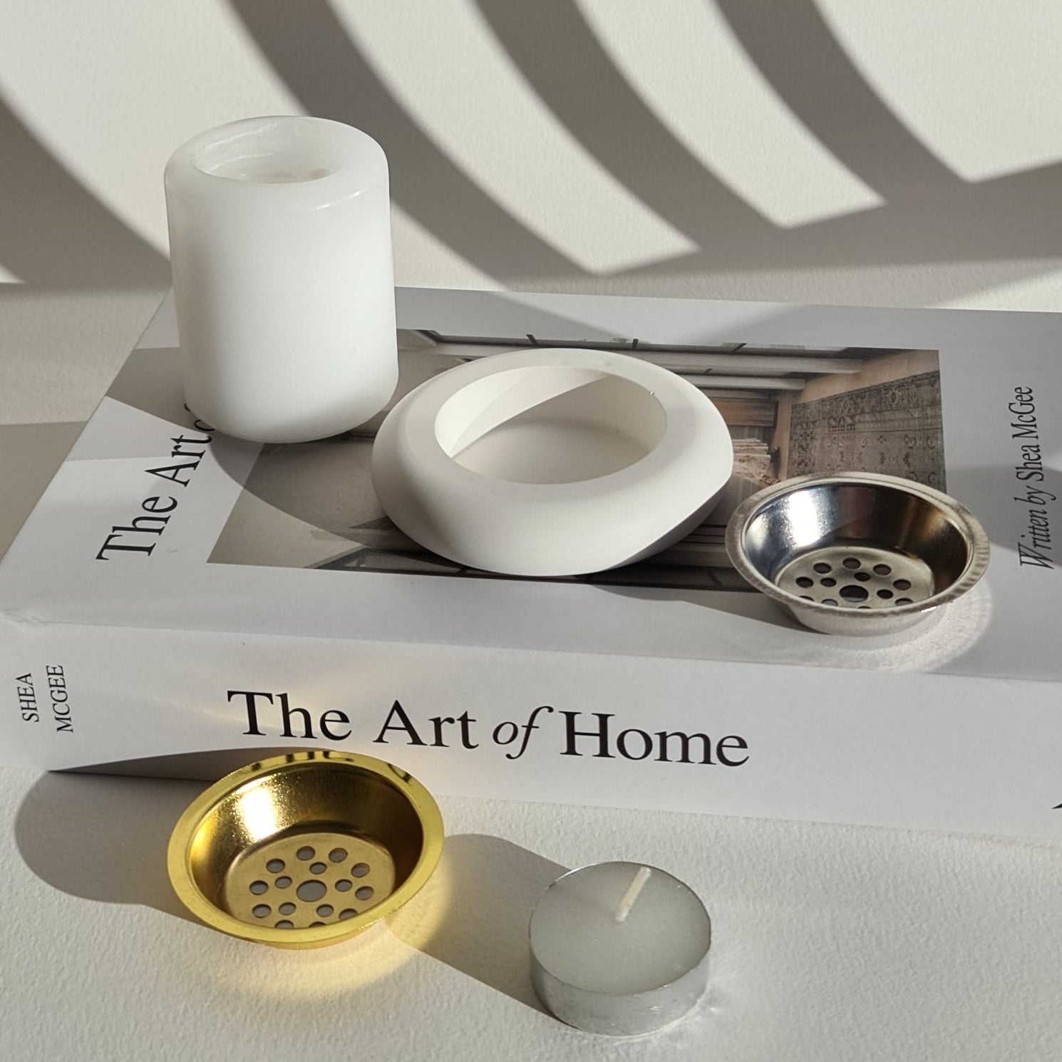 A white candle, gold and silver metal incense plates, a tealight, and a book titled 'The Art of Home' on a light surface.