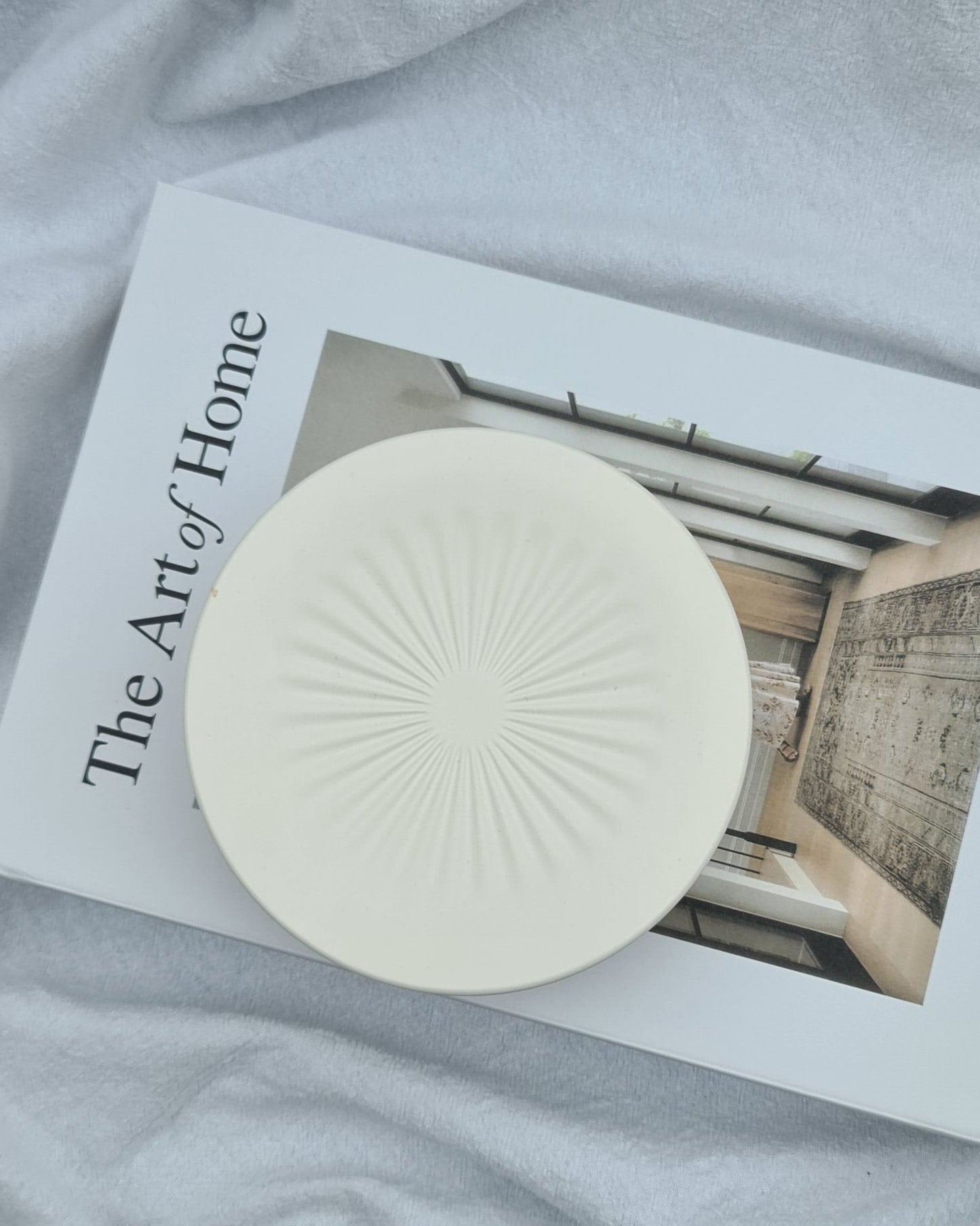 Natural white circular jar on a book titled 'The Art of Home' on a textured surface.