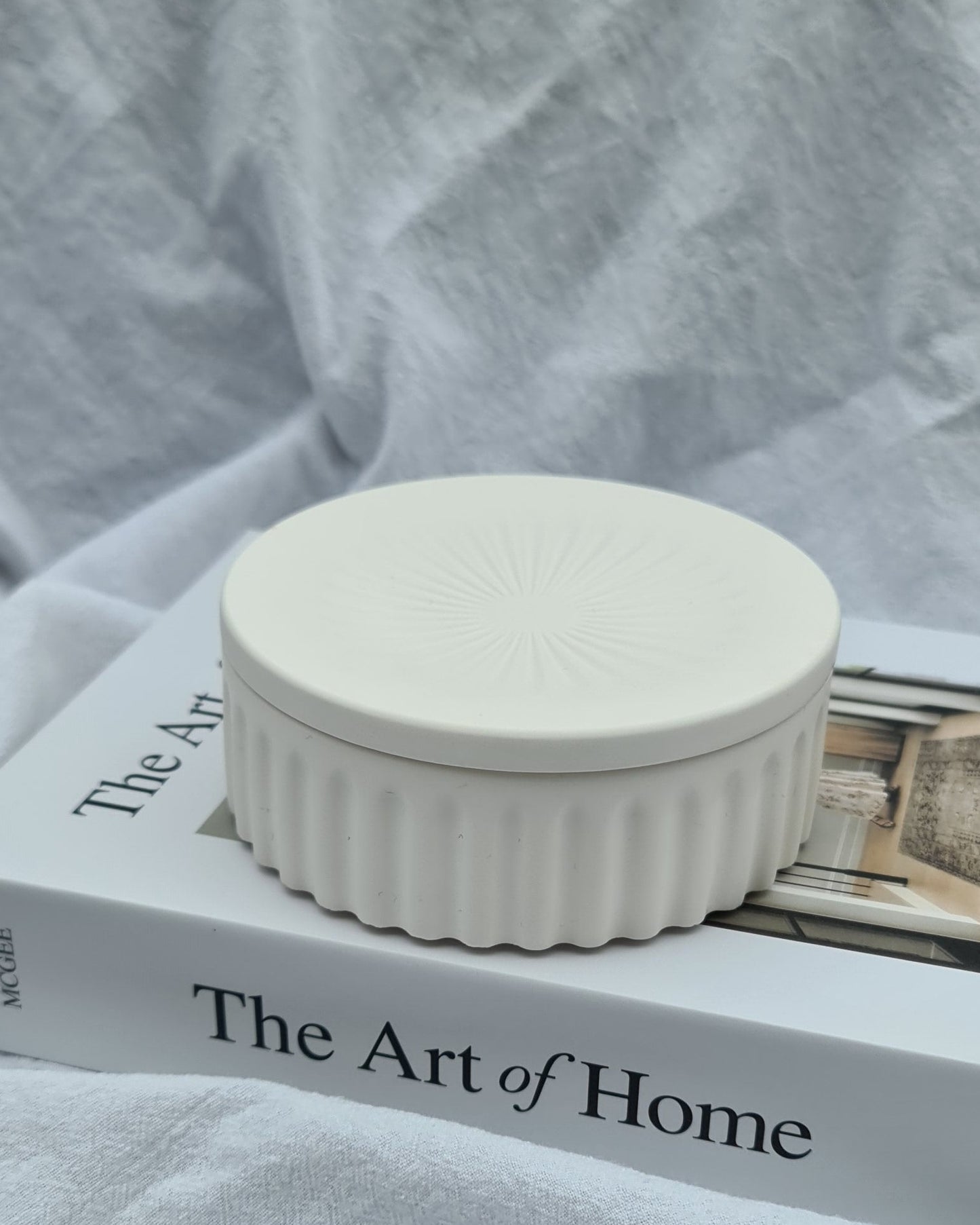 Closed natural white jar, called Wasl, on a book titled 'The Art of Home' in natural light with white fabric background.