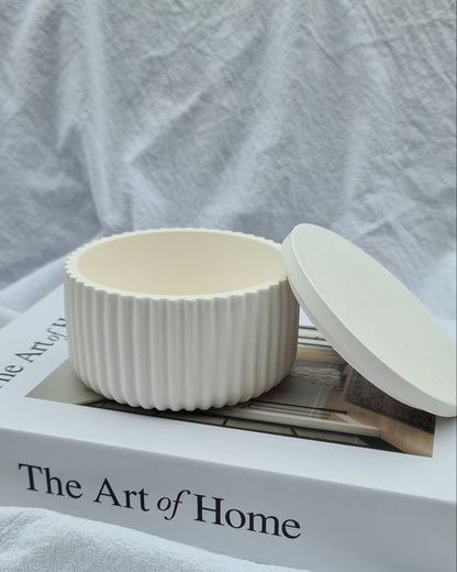 Lina wide ribbed Jar, lid leaning on side of jar, on a white linen sheet, in natural light, on a book titled 'The Art of Home'.