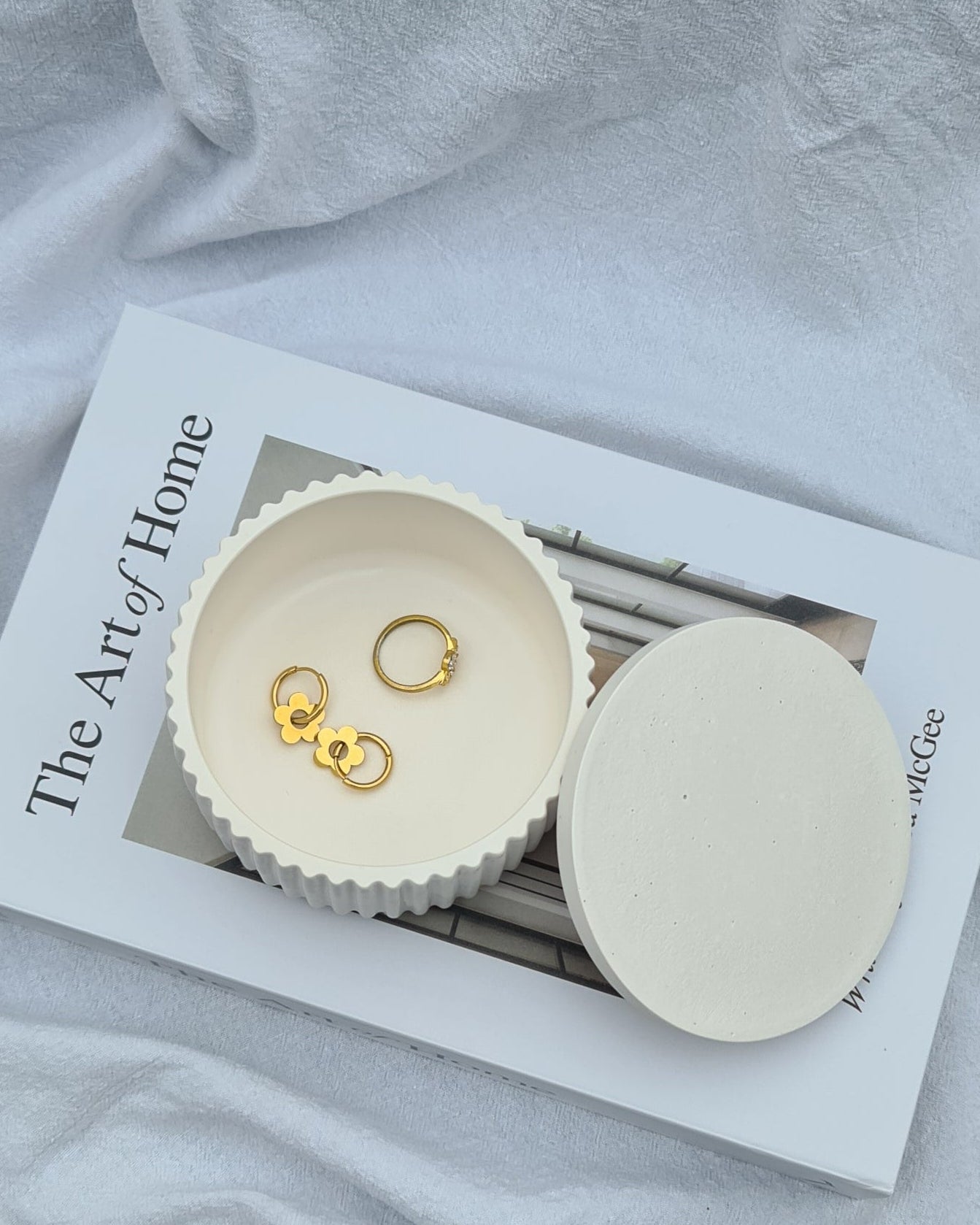 Gold earrings in the white Lina jar on a book with 'The Art of Home' on cover, in natural light