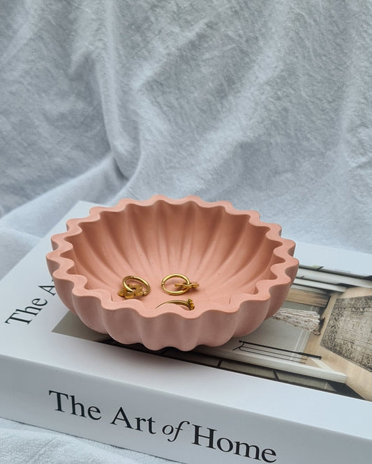 A fluted dusty rose bowl with gold earrings on a book titled 'The Art of Home' and white fabric in background