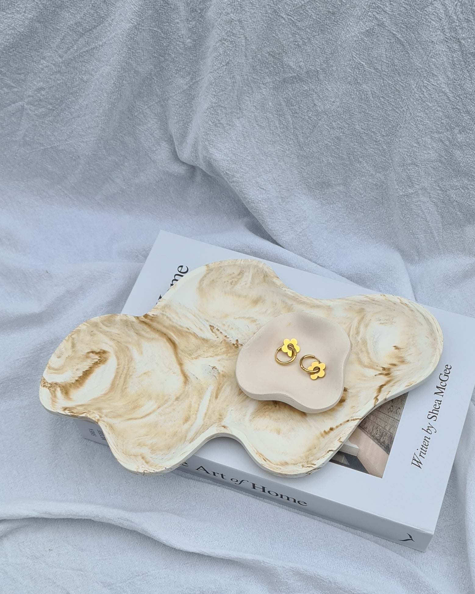 Marble-patterned jewelry Dune tray with the Petal dish with gold earrings on a book, on a white linen sheet