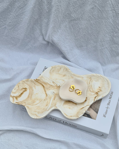 Marble-patterned jewelry Dune tray with the Petal dish with gold earrings on a book, on a white linen sheet