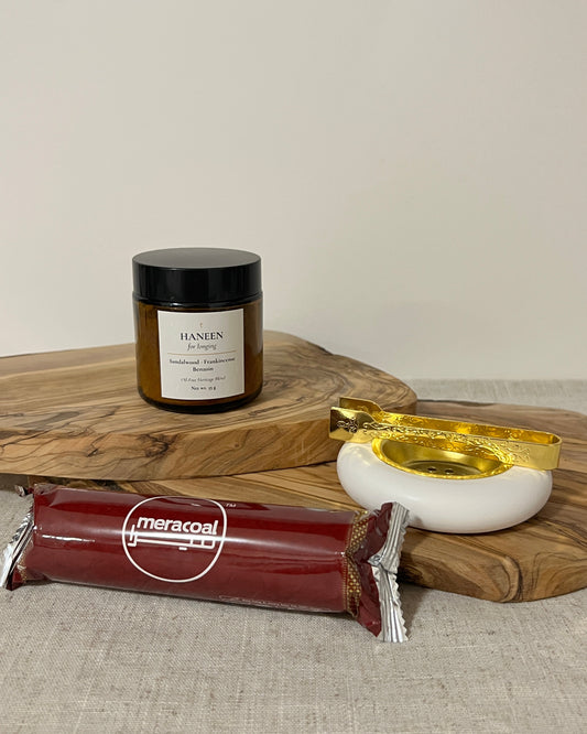 Wooden tray with an amber jar of oil-free bukhoor, Layali burner, and a pack of charcoal discs on a neutral background.