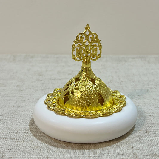 Gold metal dome-lid on white low base, this Ilaaji bukhoor burner sits on neutral fabric
