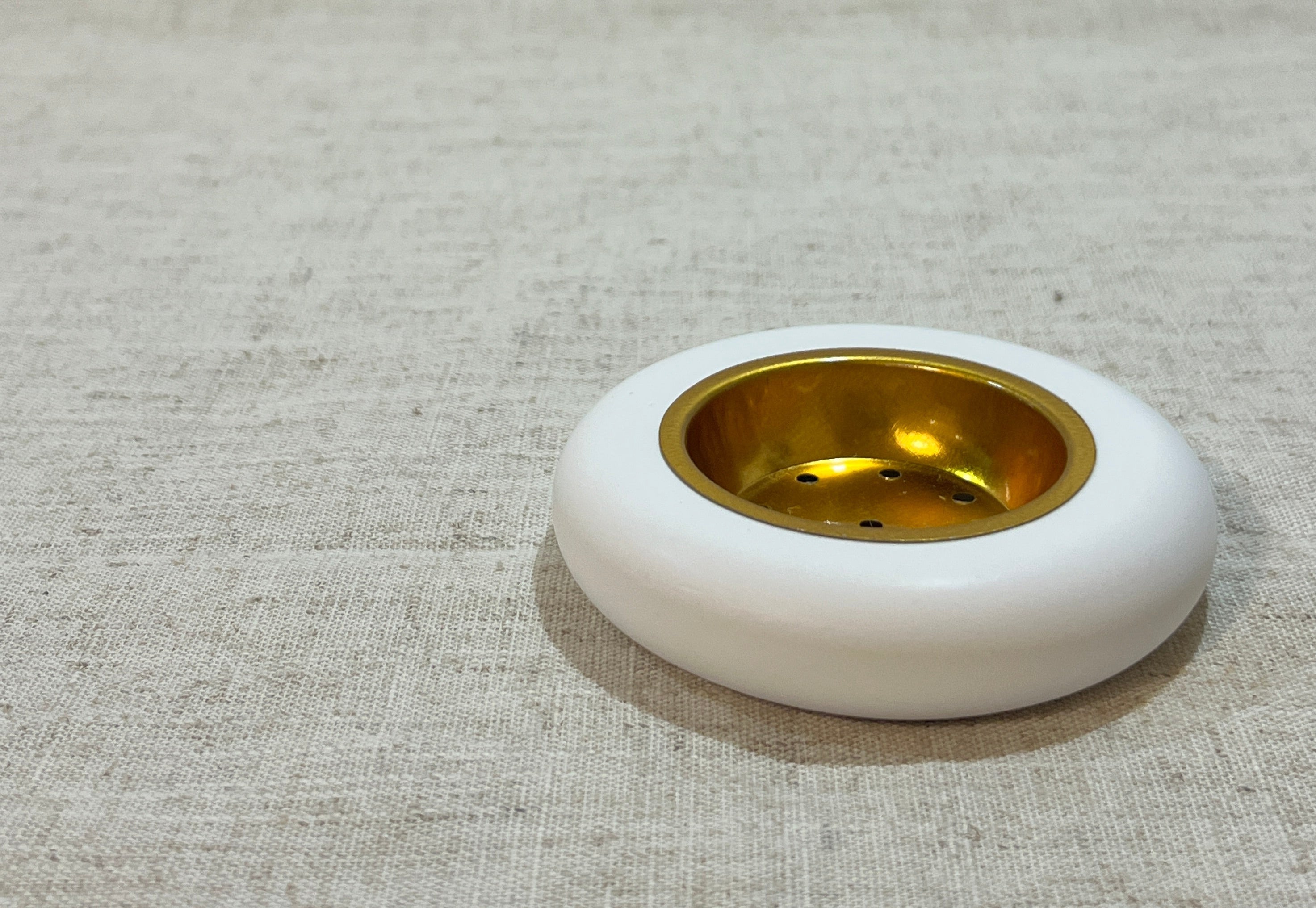 Round white bukhoor burner with gold metal plate on a textured beige .
